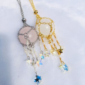 May include: Two delicate necklaces with star-shaped charms and crystal beads. One necklace features a pink quartz stone encased in a silver cage, while the other has a white stone in a gold cage.