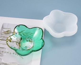 May include: A clear, flower-shaped dish with a green tint, containing gold flakes and botanical elements. Beside it, a white, flower-shaped mold. The dish is likely for decorative or functional use, possibly for jewelry or small items.