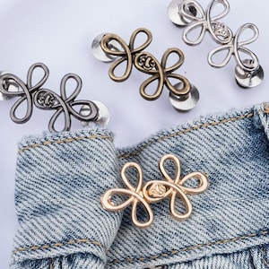 Puede incluir: Varios botones met&aacute;licos con forma de flor en acabados dorado, plateado y bronce. Los botones est&aacute;n sujetos a una tela vaquera, mostrando su dise&ntilde;o y uso potencial en ropa o accesorios.