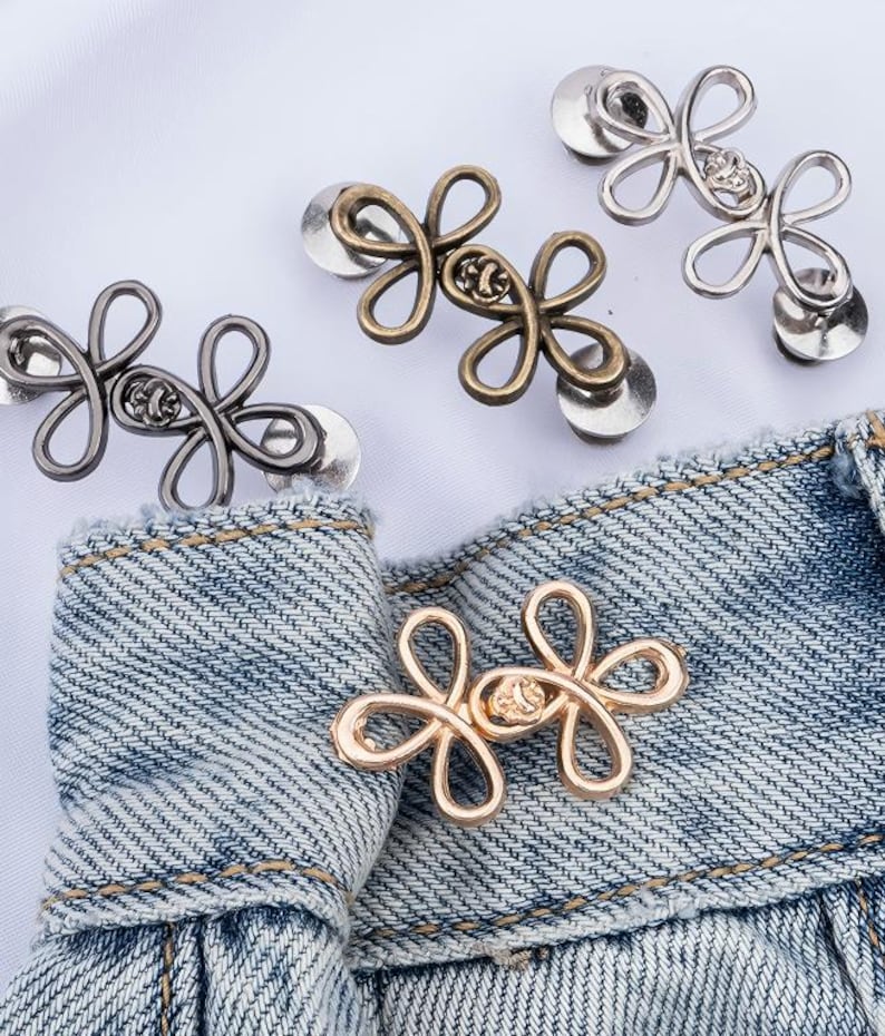 Puede incluir: Varios botones met&aacute;licos con forma de flor en acabados dorado, plateado y bronce. Los botones est&aacute;n sujetos a una tela vaquera, mostrando su dise&ntilde;o y uso potencial en ropa o accesorios.