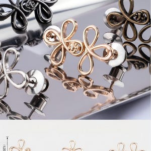 Puede incluir: Primer plano de gemelos con forma de flor en acabados plateado, dorado y ca&ntilde;&oacute;n de fusil, presentados sobre una superficie reflectante. La imagen tambi&eacute;n muestra un diagrama con medidas: 12 mm de alto y 33 mm de ancho.