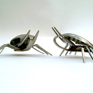 May include: Two stainless steel insect sculptures crafted from forks and spoons, set against a white backdrop. One insect faces left, the other right. Each has six legs and antennae, showcasing a unique design.