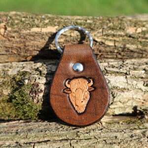 Könnte beinhalten: Ein brauner Leder-Schlüsselanhänger mit einem silbernen Schlüsselring und einem geprägten Design eines Bisonkopfes.