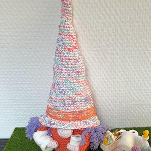 Peut inclure: Une figurine de gnome au crochet avec un grand chapeau pastel moucheté rose, bleu et blanc, orné d'une bande orange. Le gnome a un nez rond, des cheveux violets et un corps orange, tenant un petit rouleau de papier toilette blanc dans ses mains.