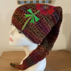 Peut inclure: Un chapeau long en crochet avec une longue queue. Le chapeau est brun, rouge et vert avec un motif de fleur et de feuille.