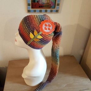 Peut inclure: Un chapeau en crochet coloré avec une longue queue. Le chapeau a un motif arc-en-ciel et une fleur en crochet avec un centre rouge et des pétales blancs. La fleur a quatre petits cœurs rouges au centre.