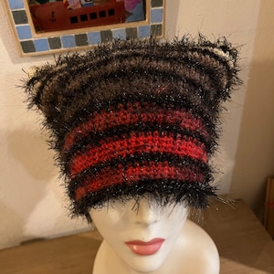 Peut inclure: Un bonnet crocheté avec une silhouette en oreilles de chat, présentant des rayures horizontales noires, marron foncé et rouges. Le fil a une finition texturée et duveteuse avec de subtils fils argentés métallisés tissés dans les sections noires.