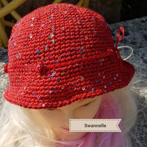 Peut inclure: Chapeau rouge en crochet avec des paillettes et un ruban rouge. Le chapeau est sur une tête de mannequin avec des cheveux blonds.