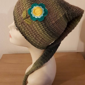 Peut inclure: Un bonnet crocheté vert et marron avec une longue queue. Le bonnet est orné d'une fleur verte avec un centre jaune et des accents bleus.