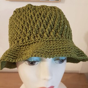 Könnte beinhalten: Ein grüner Häkel-Bucket-Hut mit breiter Krempe. Der Hut ist aus weichem, strukturiertem Garn gefertigt und hat ein schlichtes, klassisches Design.