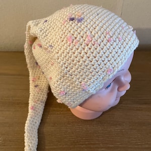 Peut inclure: Un bonnet blanc en crochet avec un long sommet pointu. Le bonnet est décoré de petites fleurs en laine rose et violette.