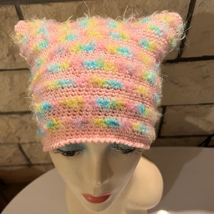 Peut inclure: Un bonnet tricoté à la main avec des oreilles de chat, présentant une palette de couleurs pastel rose clair, jaune et bleu. Le bonnet est fabriqué avec un fil texturé et duveteux qui crée un aspect doux et multicolore avec des oreilles pointues.