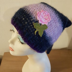 Peut inclure: Un bonnet tricoté violet et bleu avec une rose rose et des feuilles vertes brodées sur le côté.
