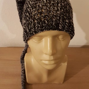 Peut inclure: Un bonnet tricoté à la main, de couleur gris foncé et blanc moucheté. Le chapeau a une longue queue pendante et est présenté sur une tête de mannequin. La texture du tricot est visible, mettant en valeur le motif.