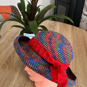 Peut inclure: Un chapeau de soleil en crochet à large bord, avec un motif rayé multicolore dans les tons de bleu, vert, rouge et rose. Le chapeau est doté d'un ruban en crochet rouge noué autour de la couronne et d'un long nœud en crochet rouge qui pend sur le côté.