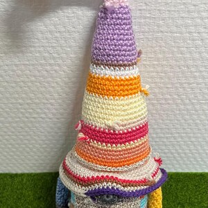 Peut inclure: Une poupée gnome crochetée à la main avec un long chapeau rose incurvé orné de pompons jaunes et roses. Le corps conique du gnome présente des rayures colorées en orange, rose et jaune. Le gnome a une barbe argentée et des bras bleus et jaunes.