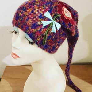 Peut inclure: Un bonnet en tricot violet, rouge et bleu avec une longue queue. Le bonnet est brodé d'une fleur et d'une libellule.