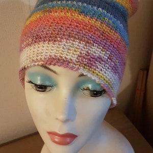 Peut inclure: Un bonnet tricoté rayé coloré avec un schéma de couleurs rose, blanc, bleu et jaune. Le bonnet est sur une tête de mannequin blanche.