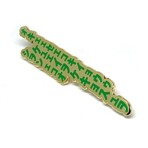 Peut inclure: Épinglette dorée avec du texte vert. Le texte est dans une langue ancienne. L'épingle est en forme de long rectangle mince.