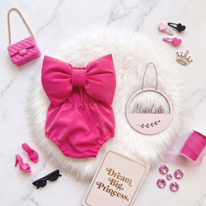 May include: A bright pink romper featuring a large bow on the chest, accompanied by a quilted pink handbag, pink high-heeled shoes, black sunglasses, heart-shaped hair clips, a crown charm, a pink ribbon, and a sign reading Dream Big, Princess.