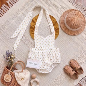 Pode incluir: Um macacão branco com bolinhas douradas, com alças de babados cor de mostarda e laços longos. Inclui um chapéu de palha, sandálias castanho-claras, uma cesta de vime com flores secas, um chocalho de madeira e uma etiqueta com Miss Lyla.