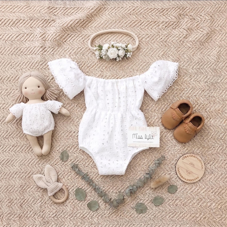 May include: A white eyelet romper with off-the-shoulder sleeves, a matching doll, a floral headband, and brown leather baby shoes. A wooden bunny teether and a wooden sign are also included. The card reads "Miss Lyla".