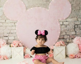 May include: A baby wearing a black off-the-shoulder top and pink polka dot shorts. The baby is also wearing a Minnie Mouse ear headband with a pink bow. A large pink Mickey Mouse head shape is in the background.