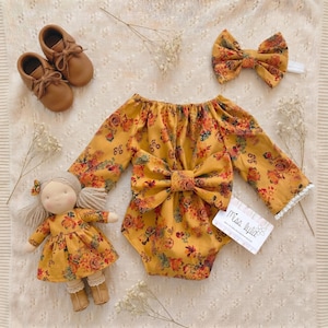 May include: A mustard yellow long-sleeve romper with a floral pattern, a large front bow, and white lace trim on the cuffs. Includes a matching floral hair bow, a pair of tan leather baby shoes, and a cloth doll wearing a matching floral dress.
