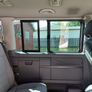 Könnte beinhalten: Innenansicht eines Vans mit einem Fenster, das einen Blick auf ein Haus und einen Zaun bietet. Der Innenraum verfügt über graue Sitze, Staufächer und eine Deckenleuchte. Das Fenster ist getönt, und die Außenansicht ist teilweise verdeckt.