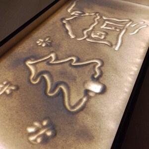 Könnte beinhalten: Eine rechteckige Sandkunstschale mit schwarzem Rahmen. Der hellbraune Sand zeigt Designs eines Hauses, eines Baumes und Schneeflocken. Die Designs sind in der Schale beleuchtet.