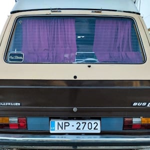 Könnte beinhalten: Rückansicht eines klassischen Volkswagen Bus D in Braun- und Beigetönen mit dem Kennzeichen NP-2702. Das Fahrzeug hat lila Vorhänge im Heckfenster. Das Wort "VOLKSWAGEN" ist sichtbar. Der Bus hat eine Retro-Ästhetik.