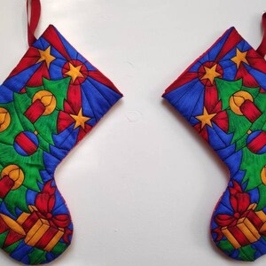 May include: Two red and blue Christmas stockings with a green Christmas tree and candles design. The stockings have a gold star and a red bow.