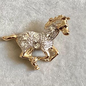 Peut inclure: Une broche en forme de cheval dorée avec des strass transparents. Le cheval est en position de course.