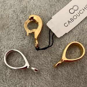 Peut inclure: Trois fermoirs de bijoux en métal doré. Deux sont des boucles simples et un est un fermoir à ressort. Les fermoirs sont sur une surface grise avec une étiquette blanche qui dit "Cabouchon".