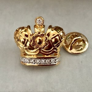 May include: A gold crown pin with clear rhinestones around the base and a single rhinestone on top. The pin has a gold back.