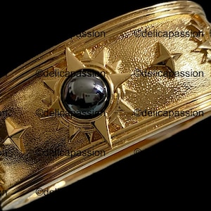 May include: A gold-colored cuff bracelet with a textured surface. The bracelet features star and sunburst designs, and is adorned with dark, spherical stones. The design is intricate and detailed, with a vintage or antique aesthetic.