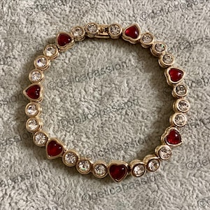Puede incluir: Pulsera dorada con piedras preciosas rojas en forma de corazón y piedras redondas transparentes alternadas. La pulsera está engarzada en una banda de metal dorado y se muestra sobre una superficie texturizada de color gris claro. Es una joya.