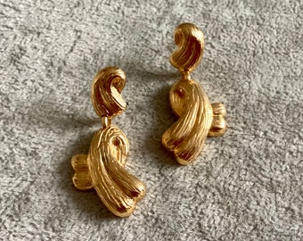 May include: A pair of gold-colored earrings with a textured, ribbon-like design. The earrings have a post and dangle style, with a curved top and a larger, looped bottom section. The earrings are displayed on a gray, textured surface.