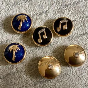 Könnte beinhalten: Eine Sammlung goldfarbener Knöpfe mit verschiedenen Designs. Einige Knöpfe zeigen eine Palme auf blauem Hintergrund, während andere eine Musiknote mit Kristallakzenten zeigen. Ein Knopf hat einen einzelnen Kristall.