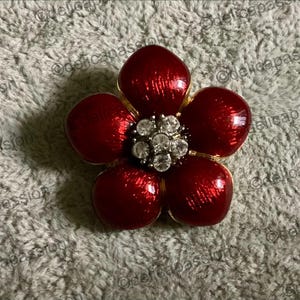 Può includere: Spilla a forma di fiore rosso con cinque petali e bordo dorato. Il centro della spilla presenta un gruppo di cristalli trasparenti e scintillanti. È un gioiello.