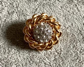 May include: A gold-toned, round brooch with a central cluster of clear rhinestones. The brooch has a braided rope design around the rhinestone cluster, creating a flower-like appearance. The background is a textured, gray fabric.