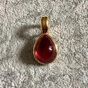 Puede incluir: Un colgante de oro con una gema roja oscura en forma de lágrima. El colgante tiene un diseño simple y elegante, con una fianza de oro para una cadena. La gema es lisa y pulida.