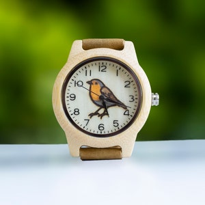 Könnte beinhalten: Eine Holzuhr mit einem braunen Lederarmband. Das Zifferblatt der Uhr hat einen weißen Hintergrund mit schwarzen römischen Ziffern als Stundenmarkierungen und eine Amsel-Illustration. Das Zifferblatt der Uhr hat auch schwarze Zeiger.