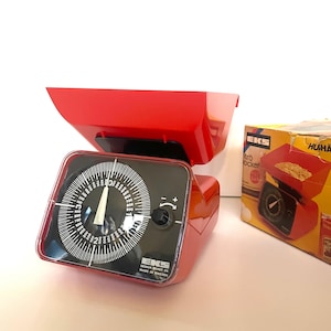May include: A red kitchen scale with a black dial and a white needle. The scale has a white label that reads "EKS Design Bengt Ek Made in Sweden".