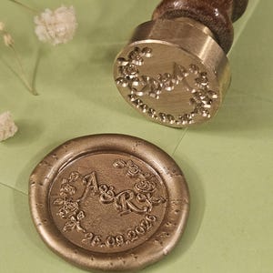 May include: A brass wax seal stamp with a wooden handle rests on a light green surface next to a wax seal. The wax seal is embossed with the initials "A&R" surrounded by floral details and the date "26.09.2020".