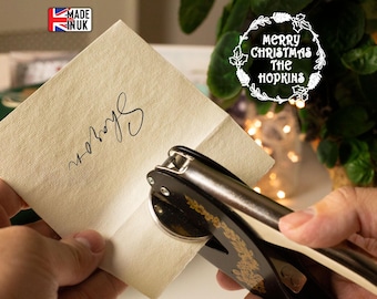 Può includere: Una persona sta usando un timbro nero e dorato per timbrare un biglietto con il testo "Merry Christmas The Hopkins". Il biglietto è beige e ha una firma a mano.