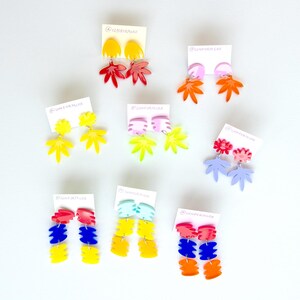 May include: A collection of colorful, abstract acrylic earrings in various shapes, including leaves and geometric designs. The earrings are displayed on white cards against a white background. The color palette includes yellow, red, blue, orange, and pink.