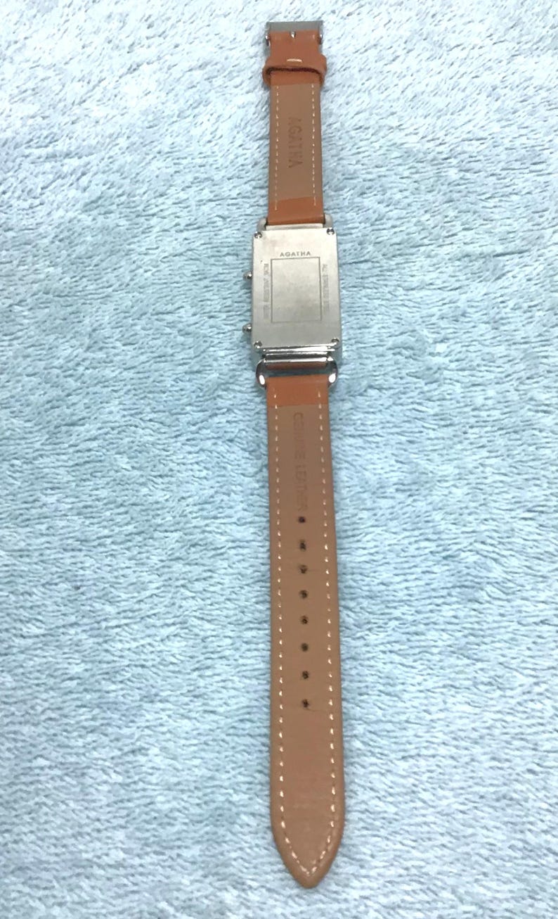 May include: A rectangular silver watch with a brown leather band. The watch face is silver, and the band is a light brown color. The watch is labeled "AGATHA" on the band and the back of the watch.