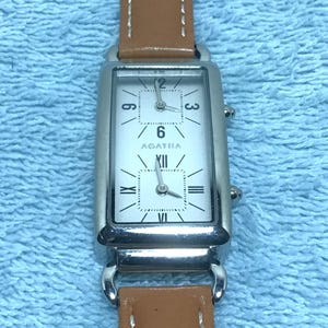 May include: A rectangular silver watch with a white face and brown leather band. The watch face displays the numbers 3, 6, 9, and 12, with the brand name "AGATHA" below the 6. The band is stitched with white thread.