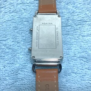 May include: A silver-toned watch with a brown leather band. The watch face has the brand name "AGATHA" and the text "WATER RESISTANT 3ATM" and "ALL STAINLESS STEEL". The band is marked "GENUINE LEATHER".